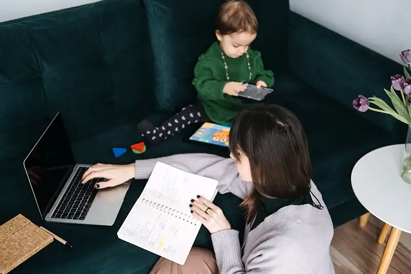 Lady trying to work using a laptop with notebook while child sits next to her. Image: irissca / Adobe Stock