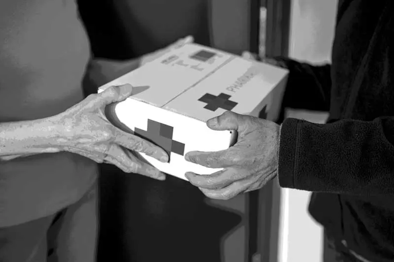 Someone handing someone else a box with a red cross on it to symbolise home prescription delivery concept. Image: vm /iStock